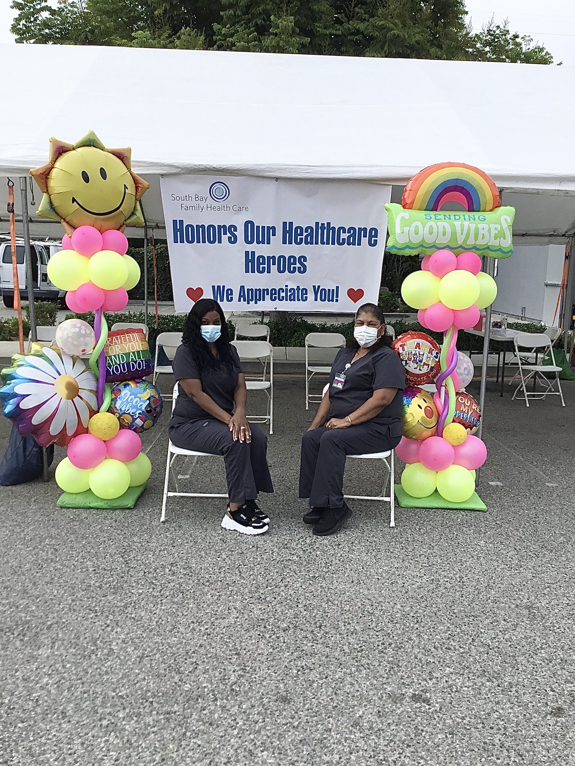 Two female medical professionals sitting in chairs under tent with sign and balloons on each side